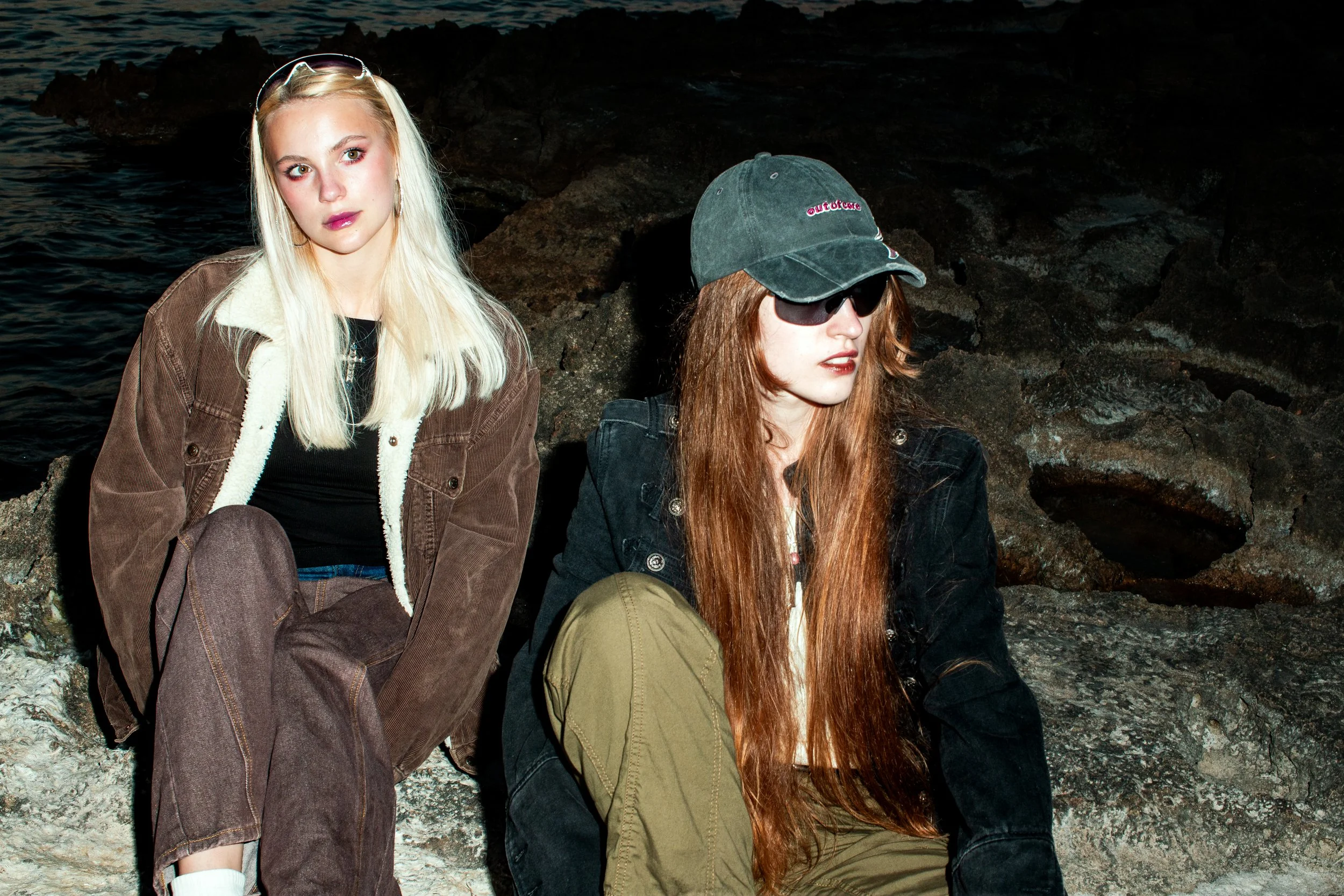 Two young women sitting on rocks by the sea at sunset, one with sunglasses and a cap, the other with long blonde hair and light eyes.