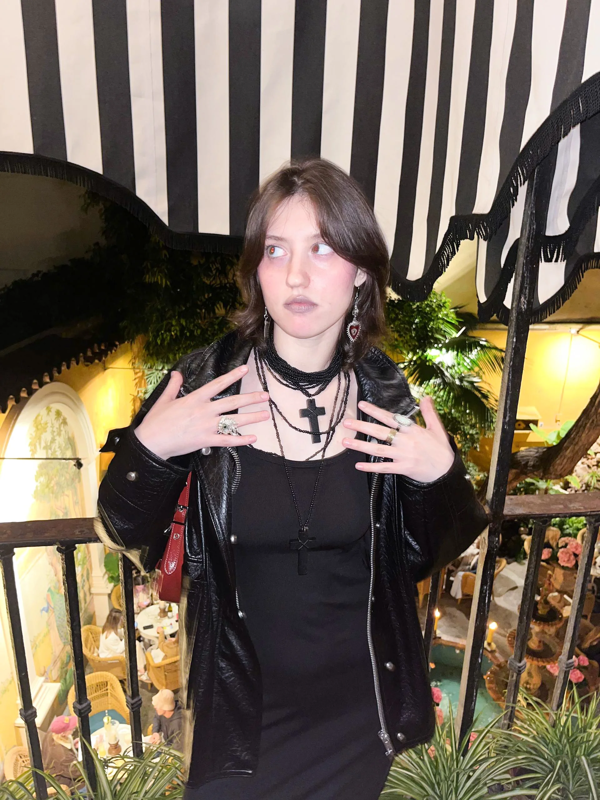 Person in black clothing with layered cross necklaces and rings posing indoors against striped curtains and lush plants