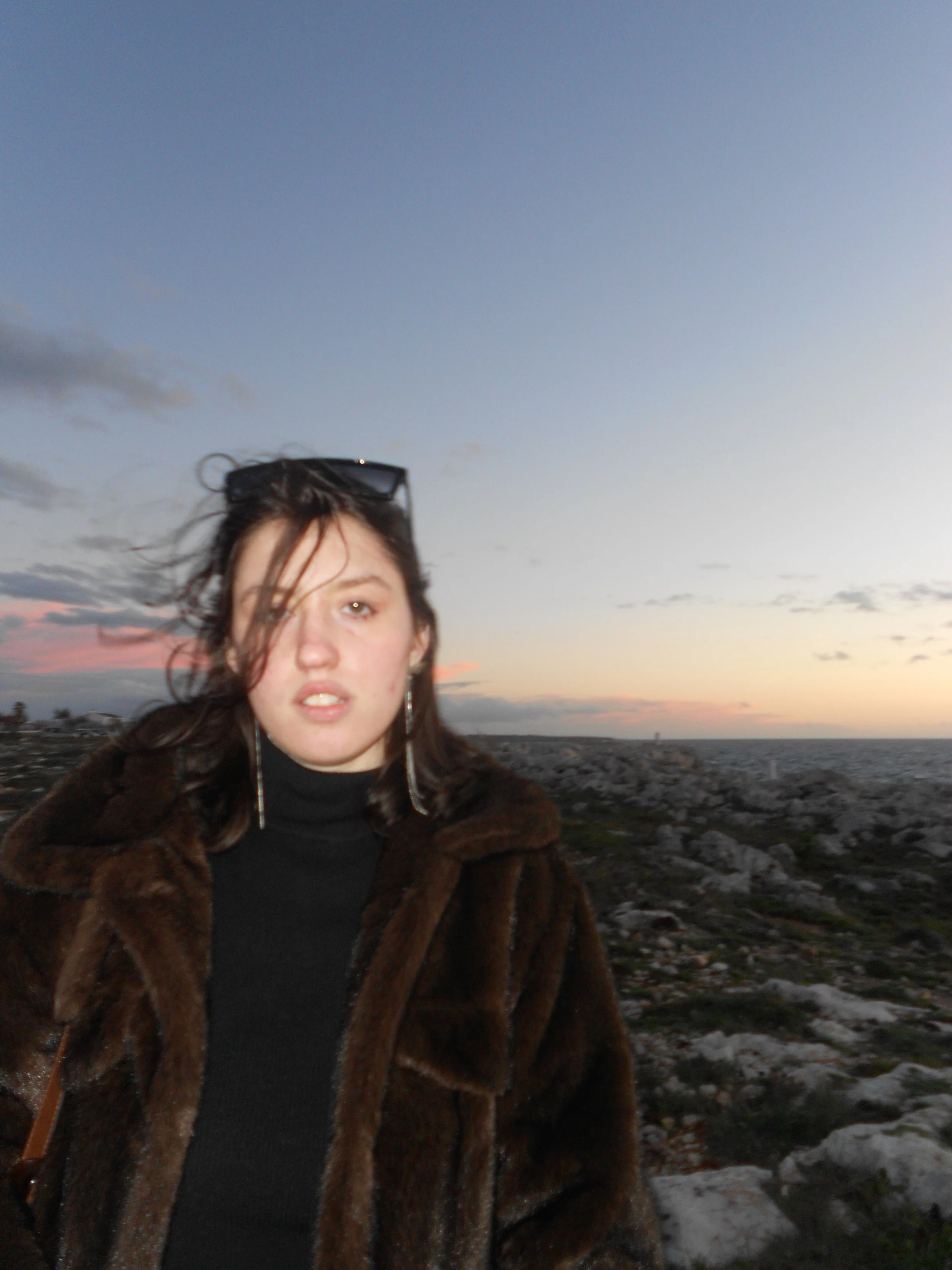 Woman in fur coat, landscape background with sunset sky and sea, dark and disheveled hair, with glasses on her head.