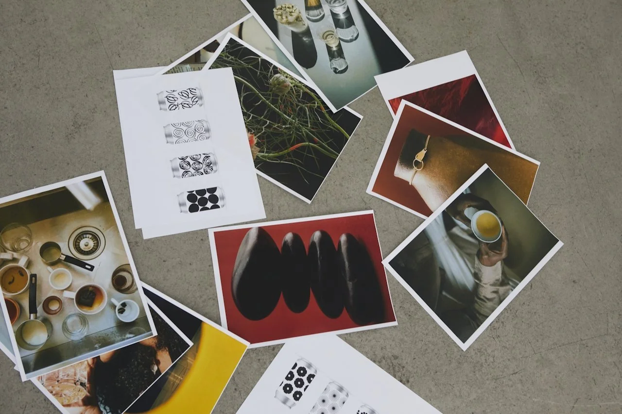 Collection of printed photographs and images spread on a concrete surface, including a top-down view of paintbrushes, abstract art, a person holding a glass of beer, and close-ups of objects and patterns.