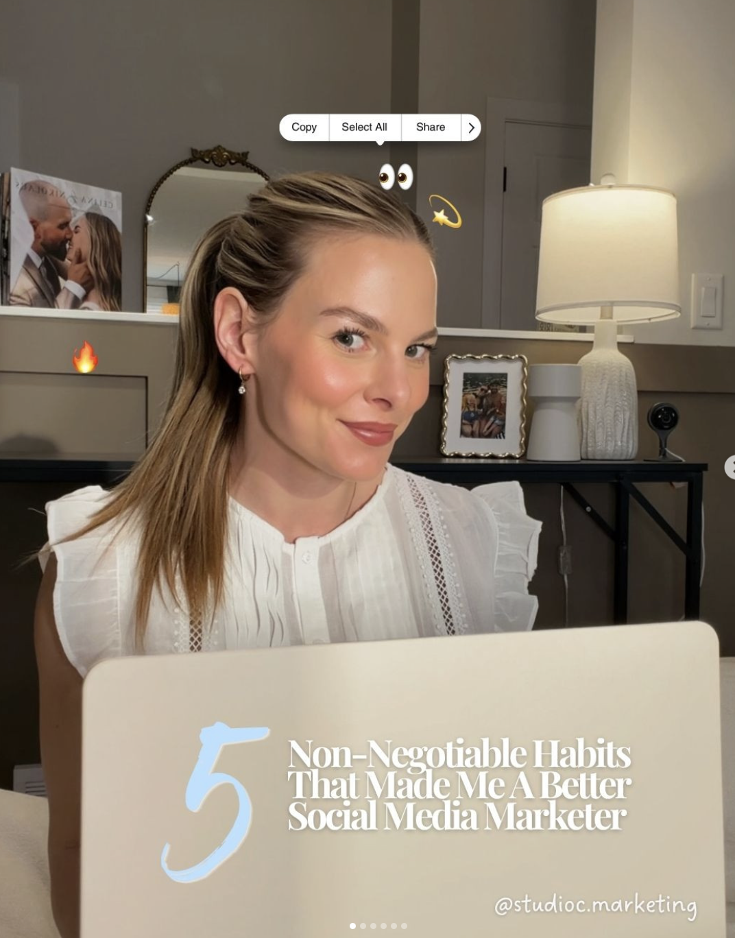 A woman with light brown hair styled in a half-up, half-down hairstyle, wearing a white blouse with eyelet details, smiling at the camera in a cozy living room. Behind her are framed photos, a mirror, a lamp, and a black table. She is holding a beige sign with the text '5 Non-Negotiable Habits That Made Me A Better Social Media Marketer' and the Instagram handle '@studioc.marketing' at the bottom.