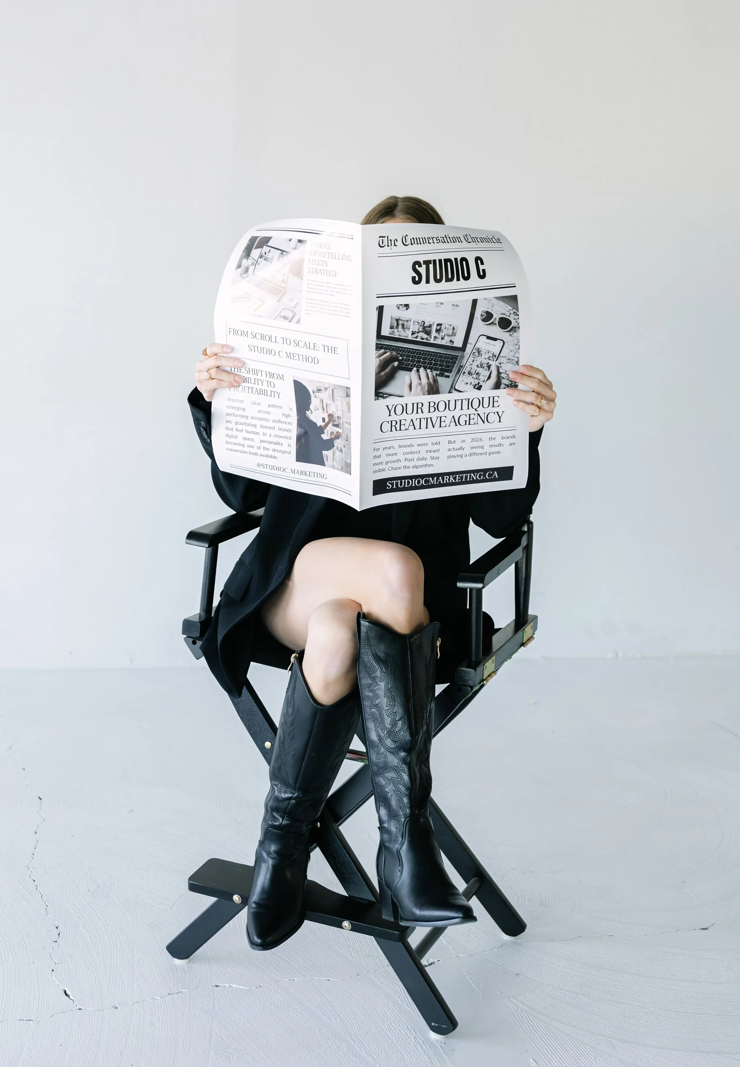 Person sitting on a director's chair, wearing black boots and a black outfit, holding a newspaper with the headline 'STUDIO C' in front of their face.