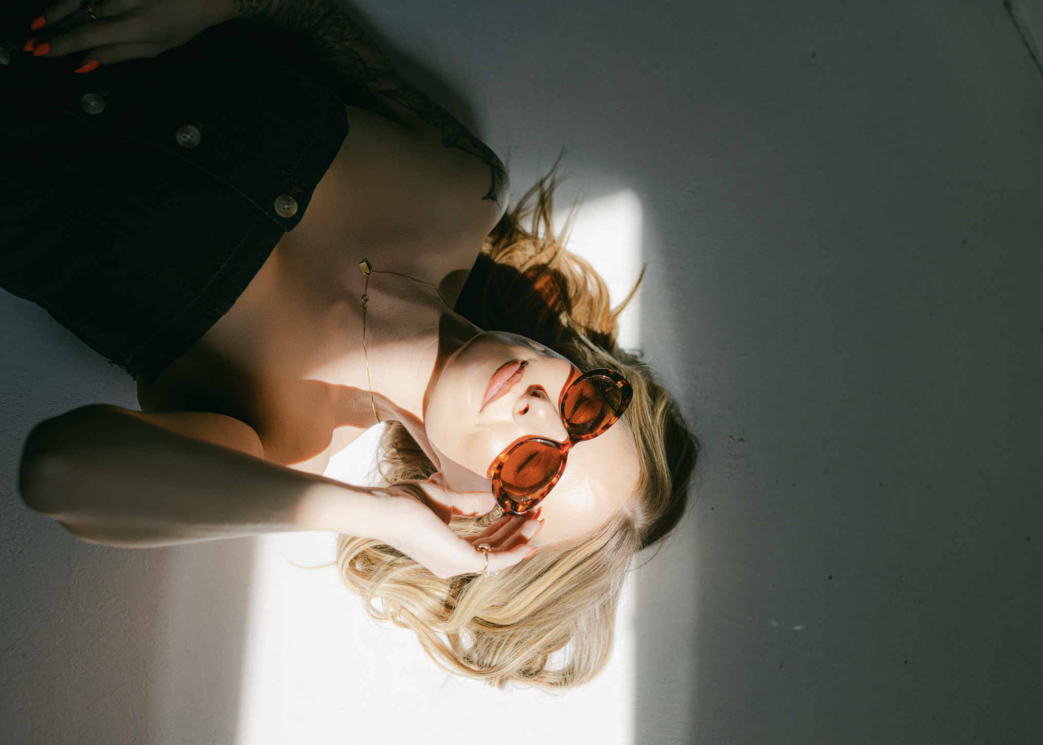 Young woman lying on the floor with sunlight casting a shadow, wearing large sunglasses, a black top, and a delicate necklace with a pendant.