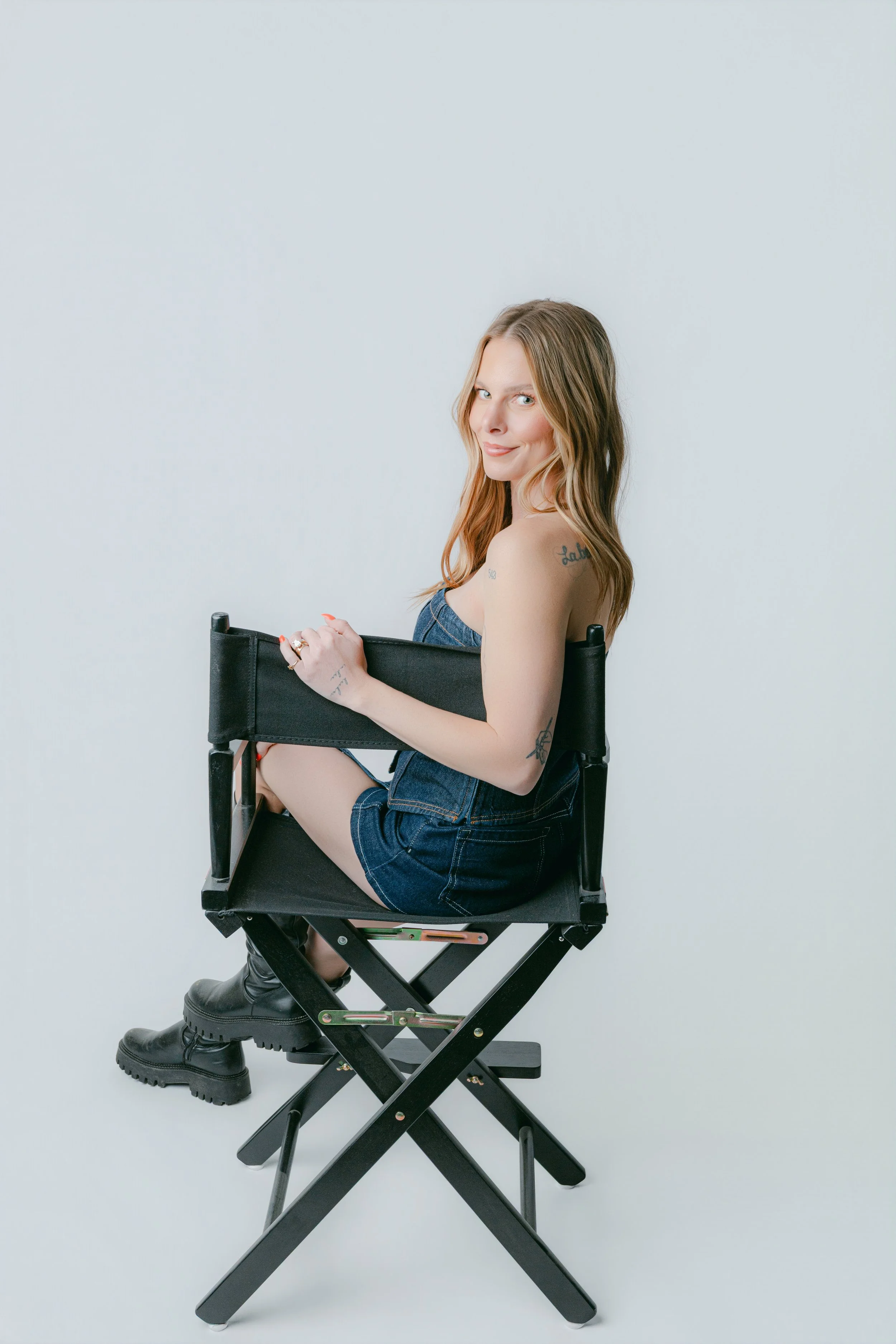 A woman with long blonde hair and tattoos, wearing a denim outfit and boots, sitting sideways on a black director's chair against a plain white background, smiling at the camera.