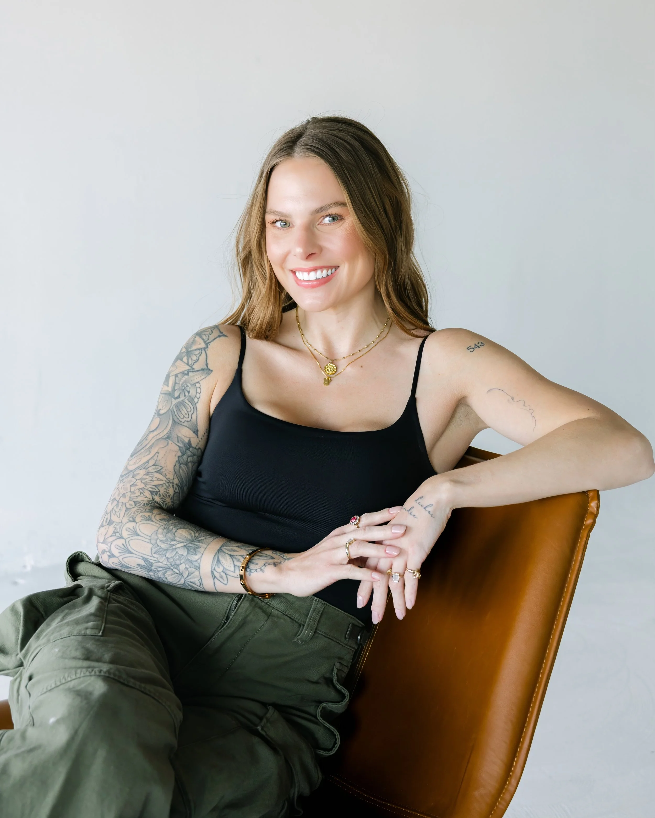 A young woman with light brown hair, blue eyes, and tattoos on her arms, smiling and taking a selfie while sitting on a brown leather chair against a plain white wall. She is wearing a black tank top with thin straps, layered gold necklaces, and rings on her fingers.