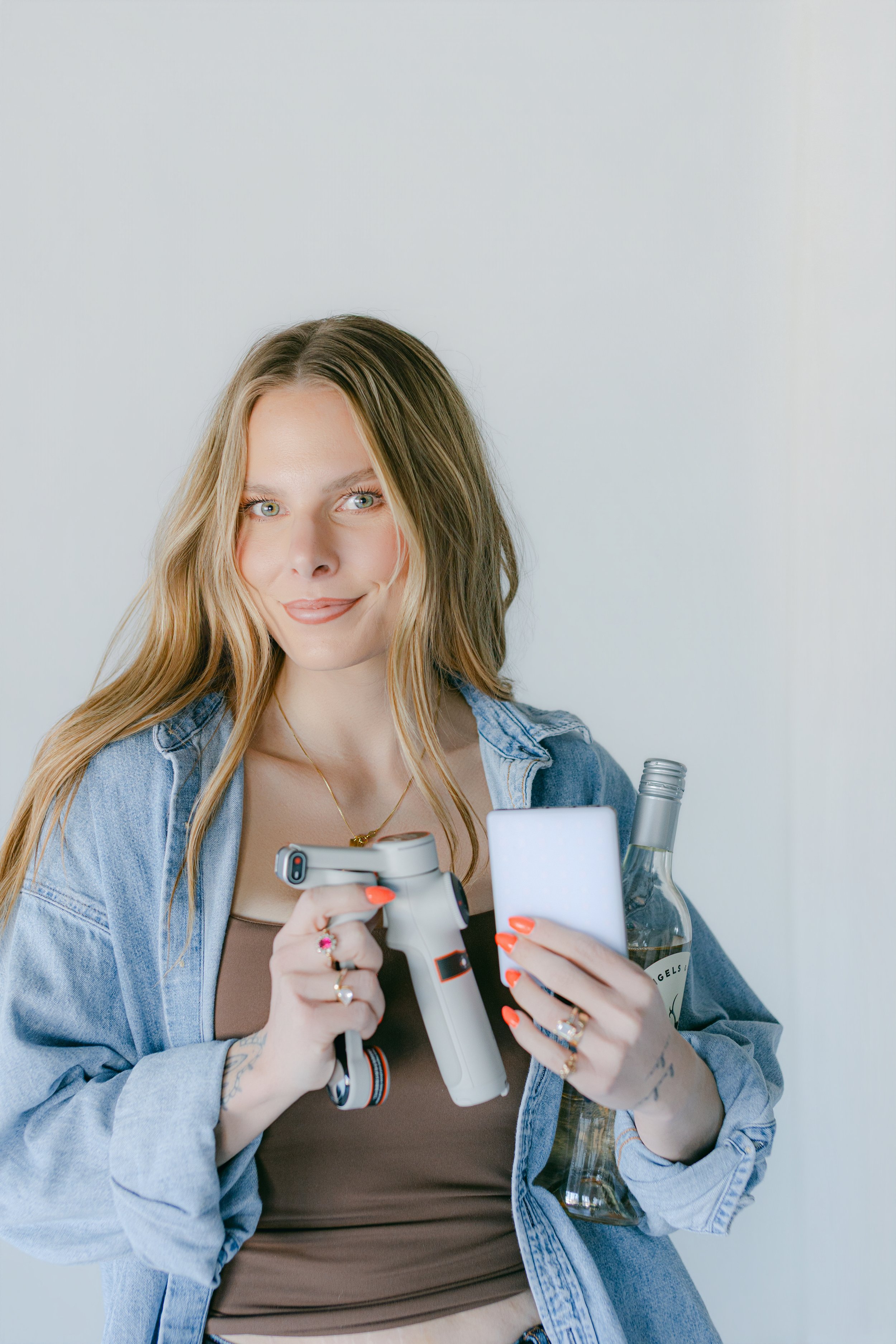 A woman with long light brown hair, blue eyes, and a light complexion wearing a denim jacket and jewelry, holding a cordless drill and a white smartphone, with a bottle of alcohol.