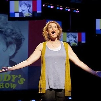 A woman, comedian Judy Gold, with curly hair and a yellow cardigan standing on stage with arms outstretched, smiling joyfully during a performance or show.