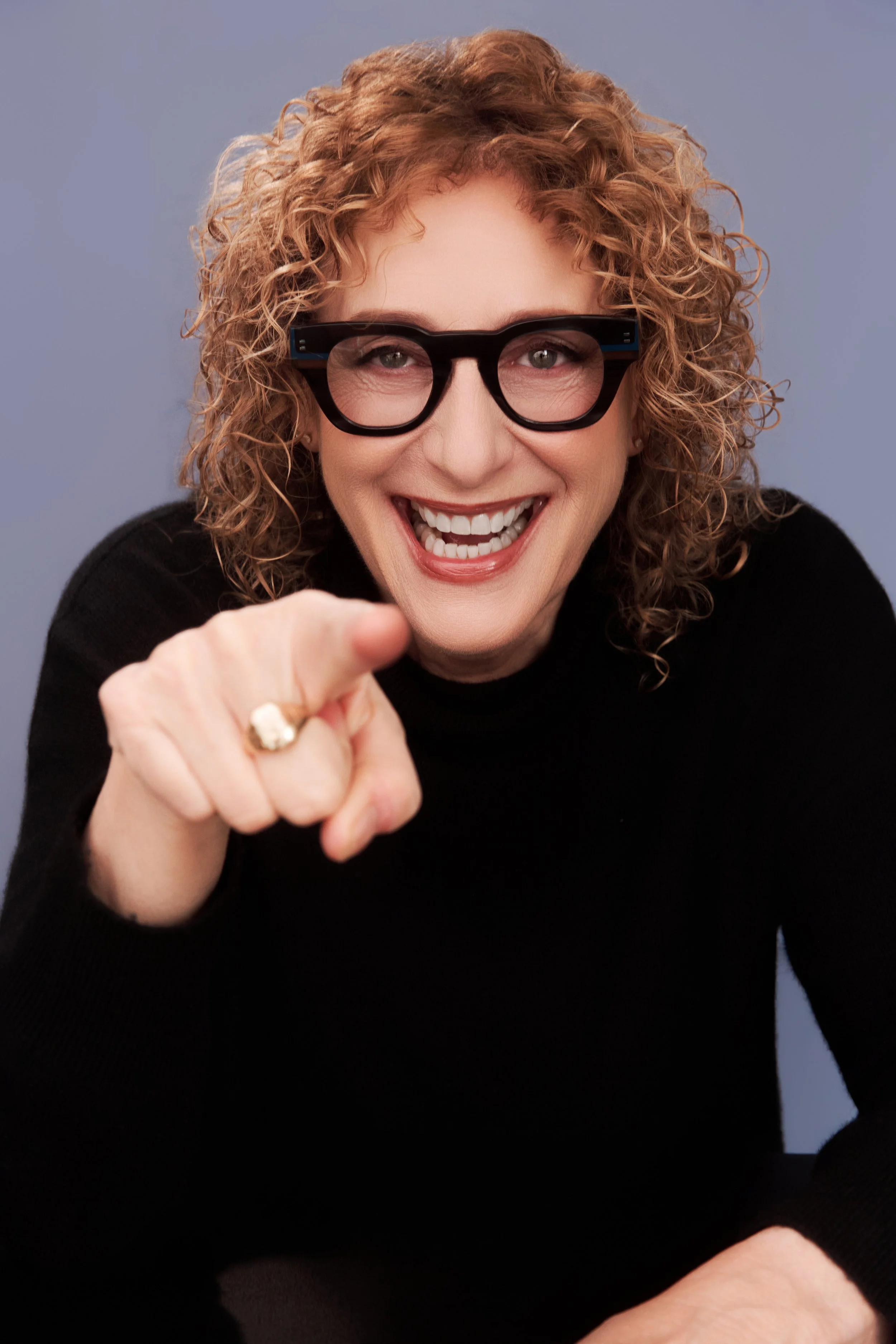 A woman, comedian Judy Gold, and black glasses smiling and pointing towards the camera, wearing a black top.