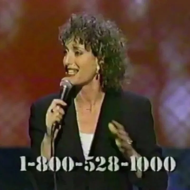 Comedian Judy Gold smiling and speaking into a microphone on stage at Comic Relief, with a phone number displayed at the bottom of the image.
