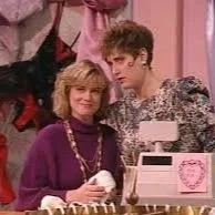 Two people, a woman with blonde hair wearing a purple sweater and a woman, comedian Judy Gold, with dark curly hair wearing a patterned shirt, standing inside a store with pink walls and a love-themed cash register.