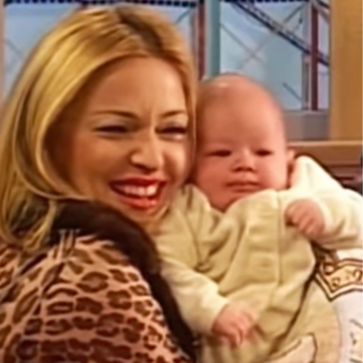 Madonna on The Rosie O'Donnell Show with blonde hair and a leopard print coat is smiling and holding a baby in a cream-colored outfit, indoors near a window with a construction crane visible outside.