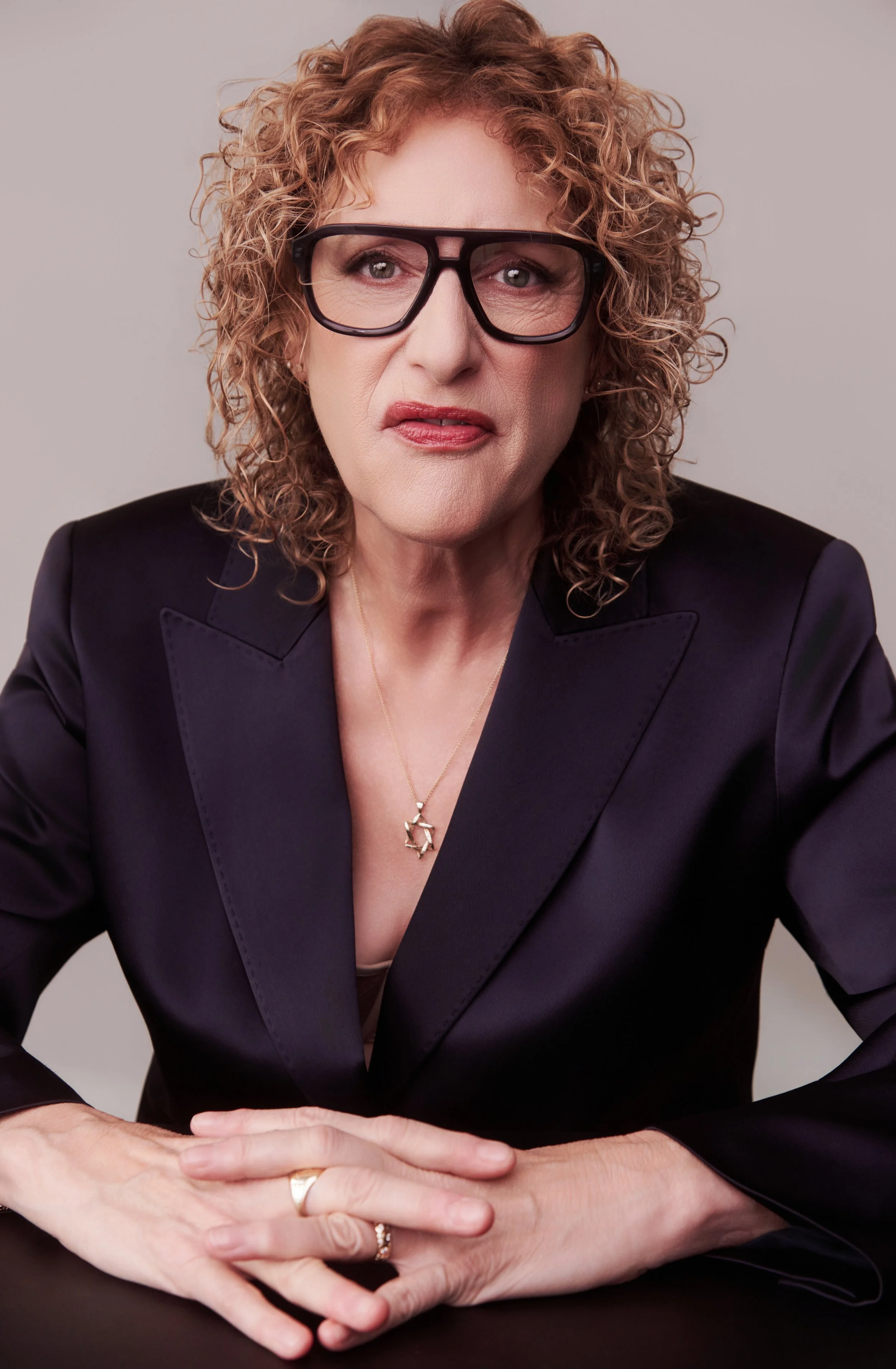 A woman with curly red hair and glasses, wearing a dark blazer with a deep neckline, sitting at a table with her hands folded in front of her.