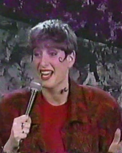 Comedian Judy Gold with short hair, wearing a red top and a maroon jacket, smiling while holding a microphone, on a stage with a stone background.