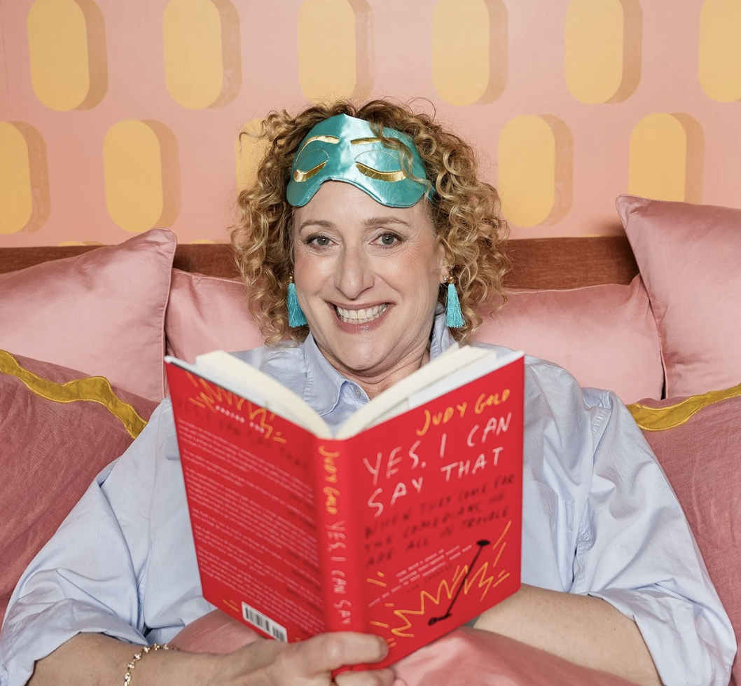 A woman, comedian Judy Gold, blue sleep mask on her forehead, and turquoise earrings, is sitting on a pink bed with pink pillows, smiling while reading a bright red book with yellow handwritten text.