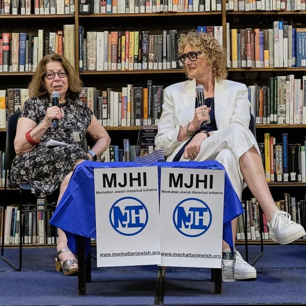 Two women seated at a table with microphones, speaking at a Manhattan Jewish Historical Initiative event, with a bookshelf background.