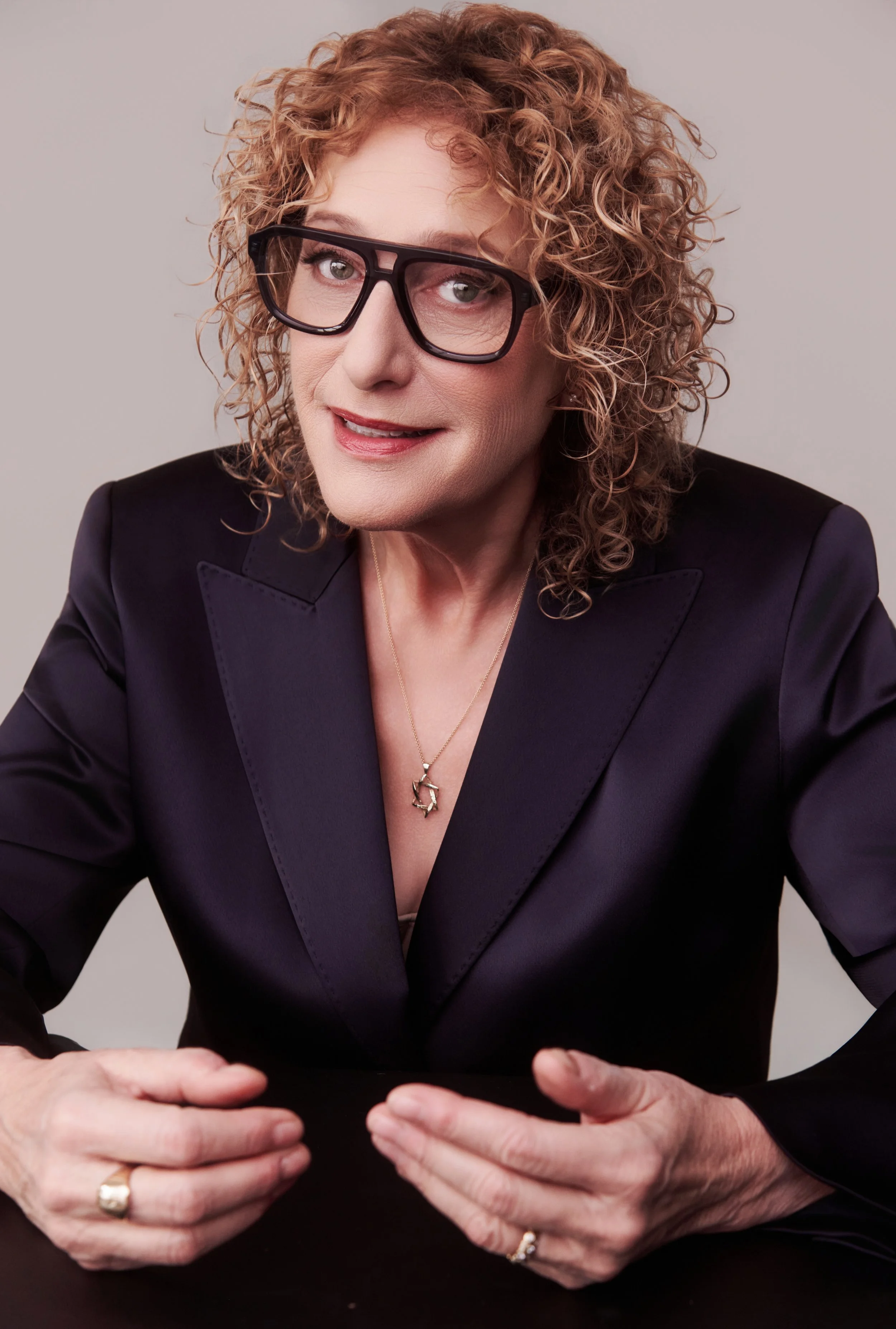 A woman, comedian Judy Gold, wearing glasses and a black blazer, sitting at a black table with her hands resting on it, against a light gray background.