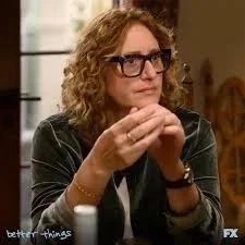 A woman, comedian Judy Gold, with curly hair and glasses sitting at a table, clasping her hands together and looking thoughtfully.