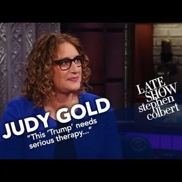 Judy Gold on the Stephen Colbert late show, wearing glasses and a blue top, with text overlay about her needing serious therapy.