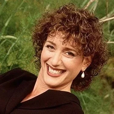 Comedian Judy Gold smiling outdoors with greenery in the background.