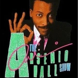 A man making an 'OK' gesture with his right hand, wearing a dark suit, with text reading 'The Arsenio Hall Show' in colorful letters.