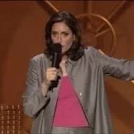 A woman, comedian Judy Gold, on stage holding a microphone and gesturing with her arm.