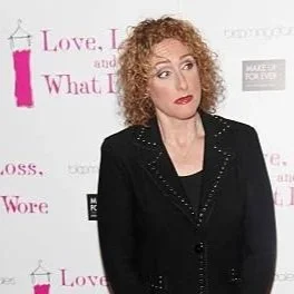 Comedian Judy Gold with curly red hair wearing a black blazer standing in front of a pink and white backdrop at a promotional event for the play Love, Loss, and What I Wore.