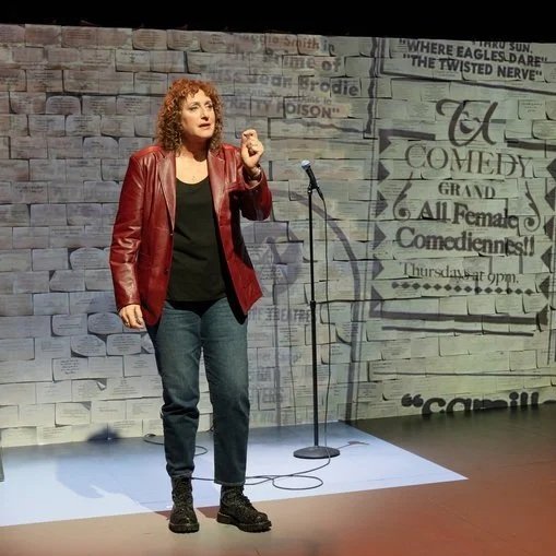 A woman, comedian Judy Gold, standing on stage, speaking into a microphone, with a brick wall backdrop and event poster projected on the wall.