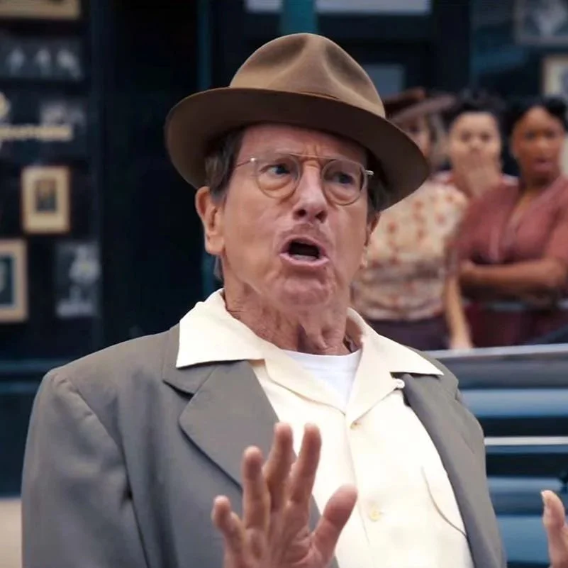 Larry David wearing a brown hat, glasses, and a gray blazer is speaking passionately with his hand raised. In the background, there are three women watching with concerned and surprised expressions.