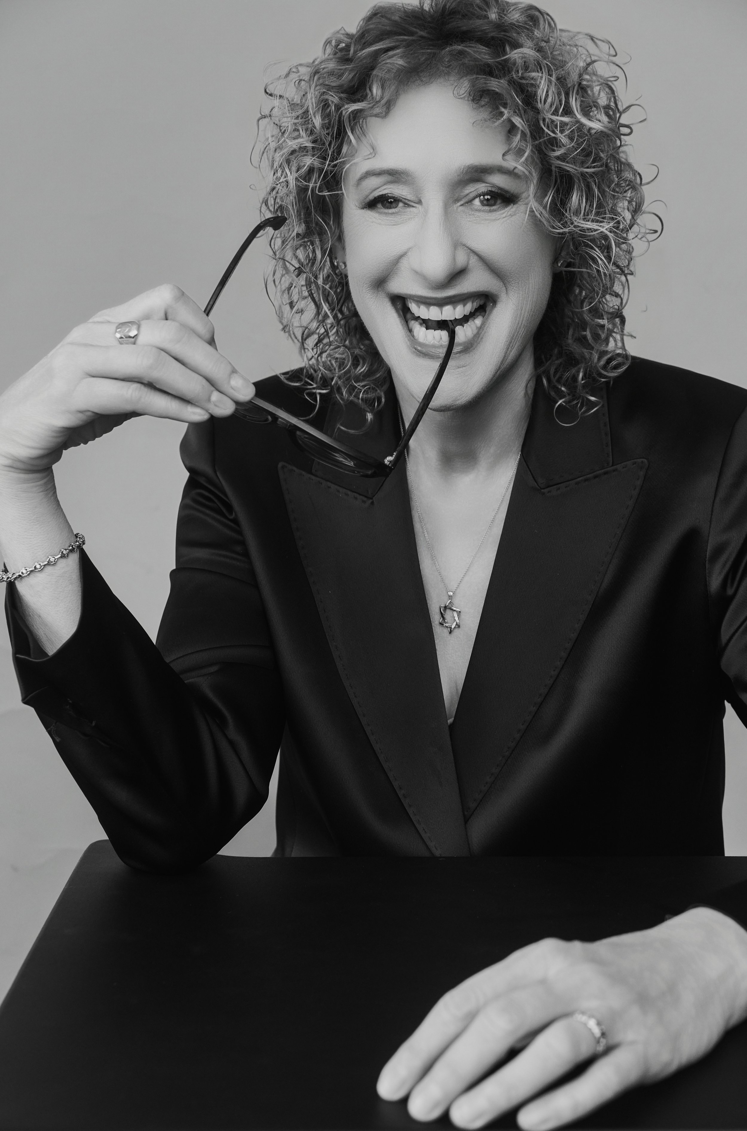 Black and white photo of a woman, comedian Judy Gold, holding sunglasses near her mouth, showing a playful expression, wearing a dark blazer and a star-shaped necklace, with her hand resting on a table in front of her.