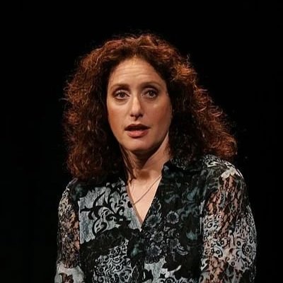 A woman, comedian Judy Gold, with curly brown hair wearing a black and white patterned blouse, against a dark background.