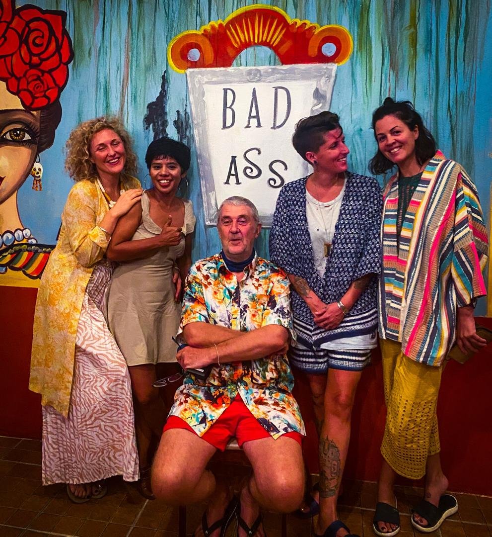 Five people smiling and posing in front of a colorful mural with the words "BAD ASS." One person is sitting with arms crossed, wearing a floral shirt and red shorts, while the other four are standing next to him, all dressed casually and smiling.