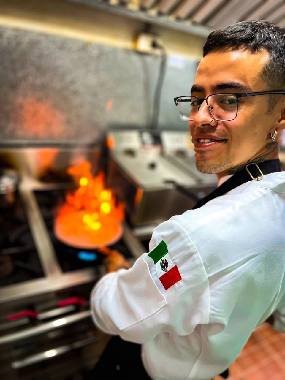 A male chef wearing glasses and safety earrings, smiling while cooking on a stove. He is wearing a white chef's jacket with a Mexican flag patch on the sleeve, and he is stirring a pan with what appears to be flames from the burner visible.