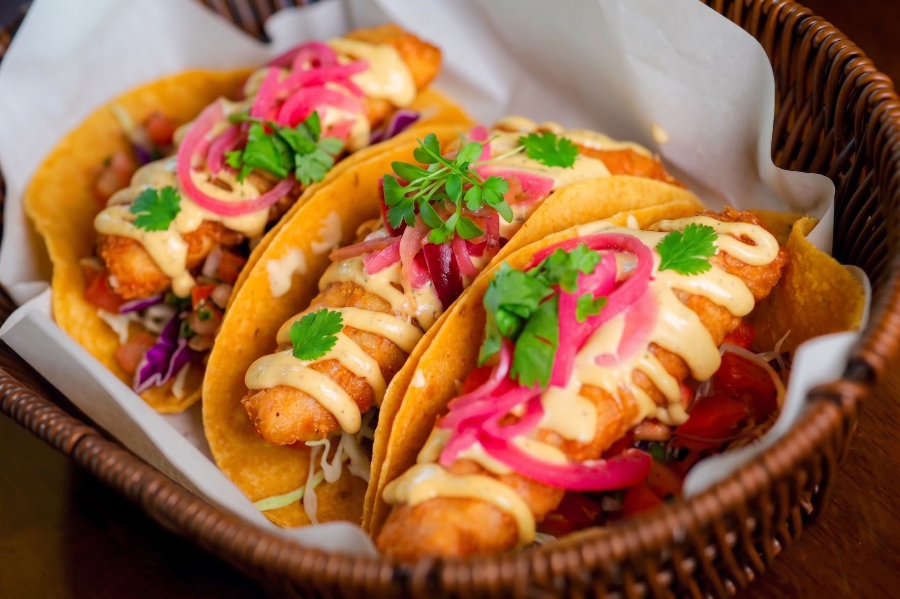 Three fish tacos in a basket garnished with cilantro and pickled red onions, topped with drizzled sauce.