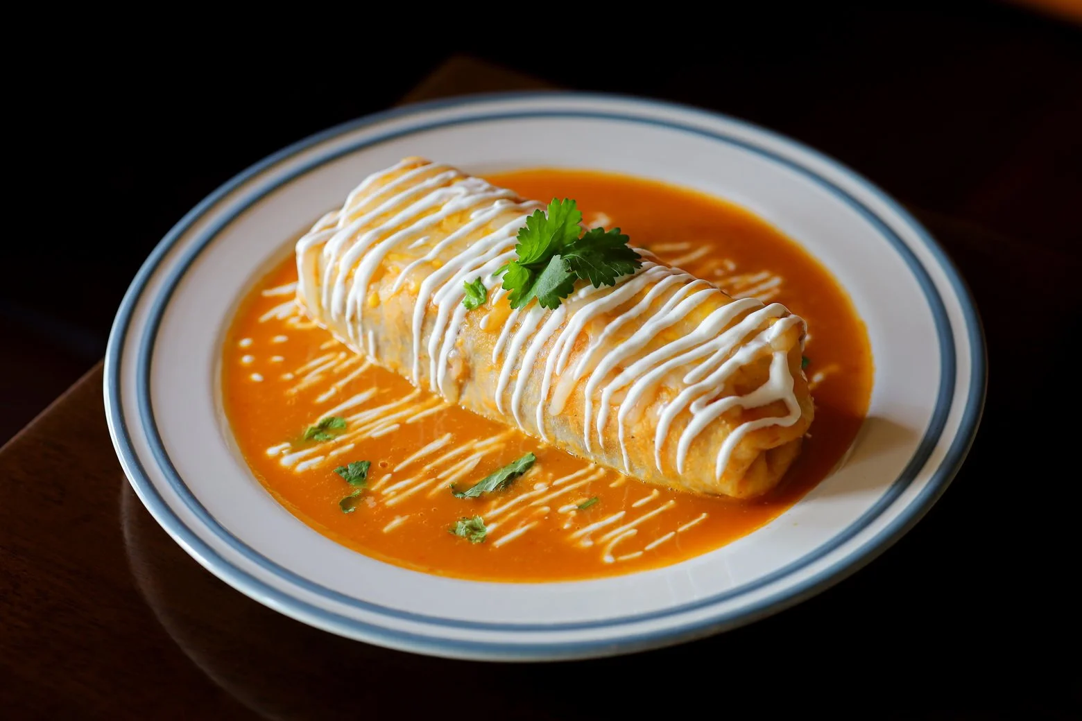A plate of Mexican cheese enchilada topped with white sauce and garnished with cilantro, served in orange enchilada sauce.