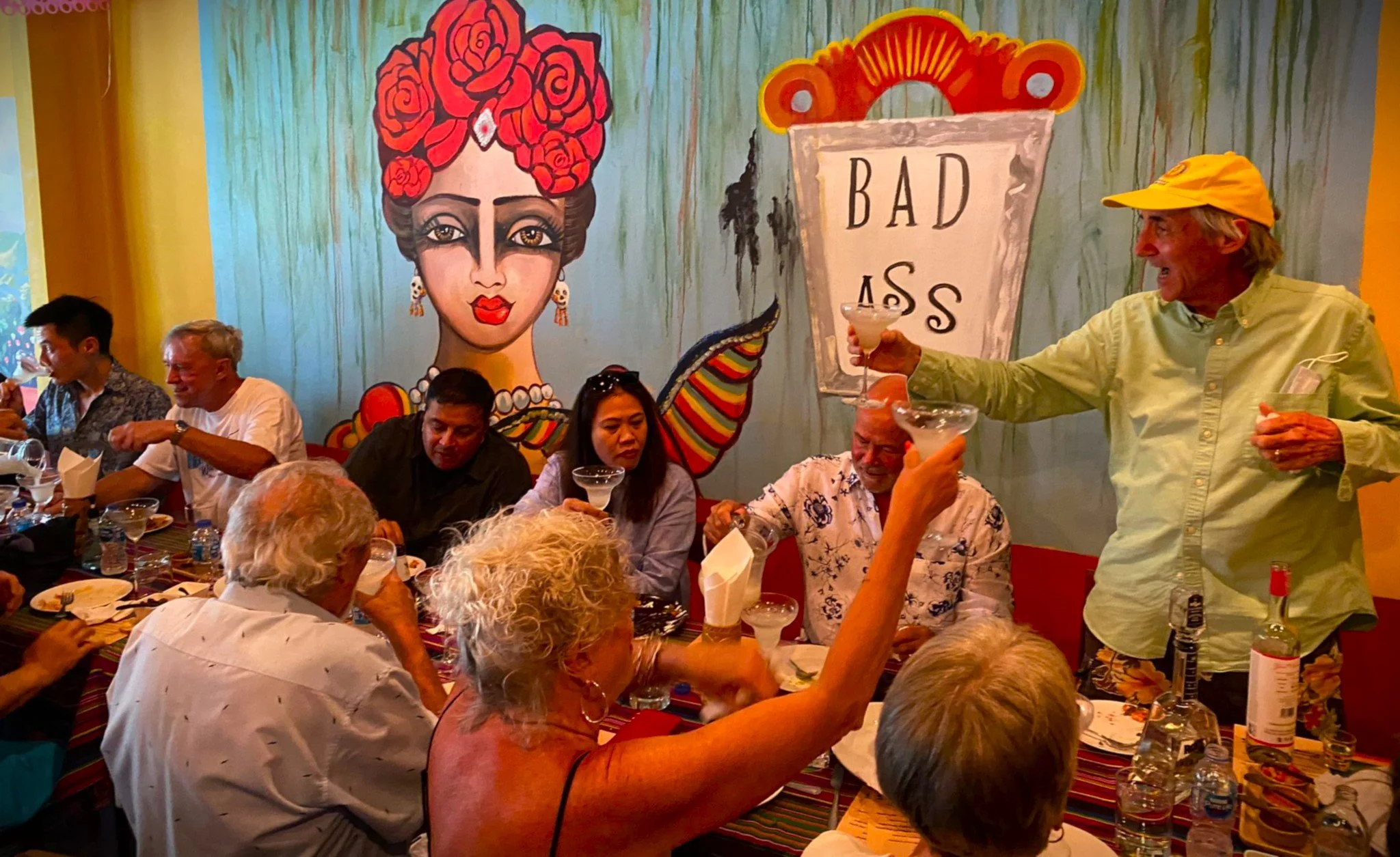 A lively restaurant scene with people seated at a long table, enjoying drinks and conversation. The background features a colorful mural of a woman with red roses in her hair and a sign that says 'Bad Ass.' A man in a yellow shirt and cap is standing