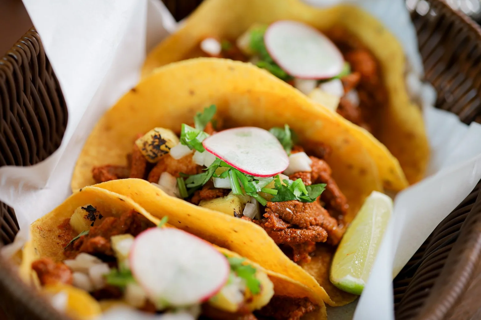 Three tacos in a basket with lime wedges, topped with radish slices, onions, and cilantro.