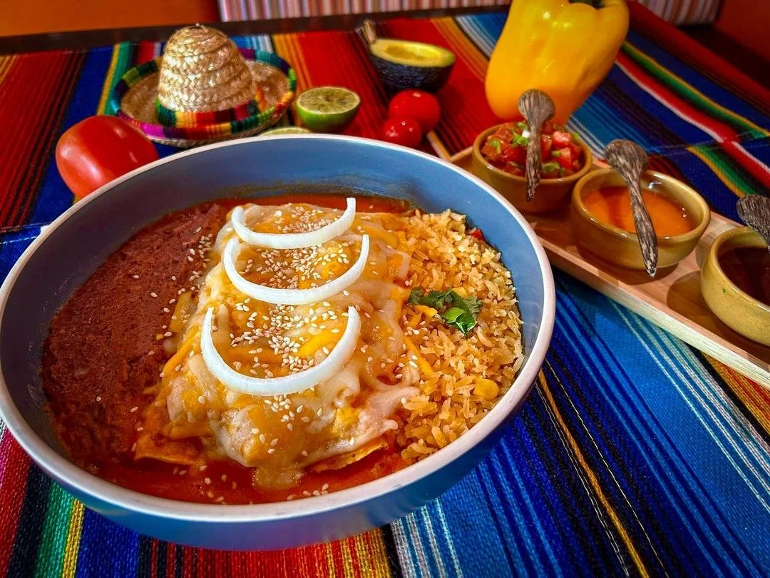 Bowl of Mexican food with ground beef, melted cheese, sour cream, rice, and beans, surrounded by salsa, lime, bell pepper, tomatoes, and a small sombrero.