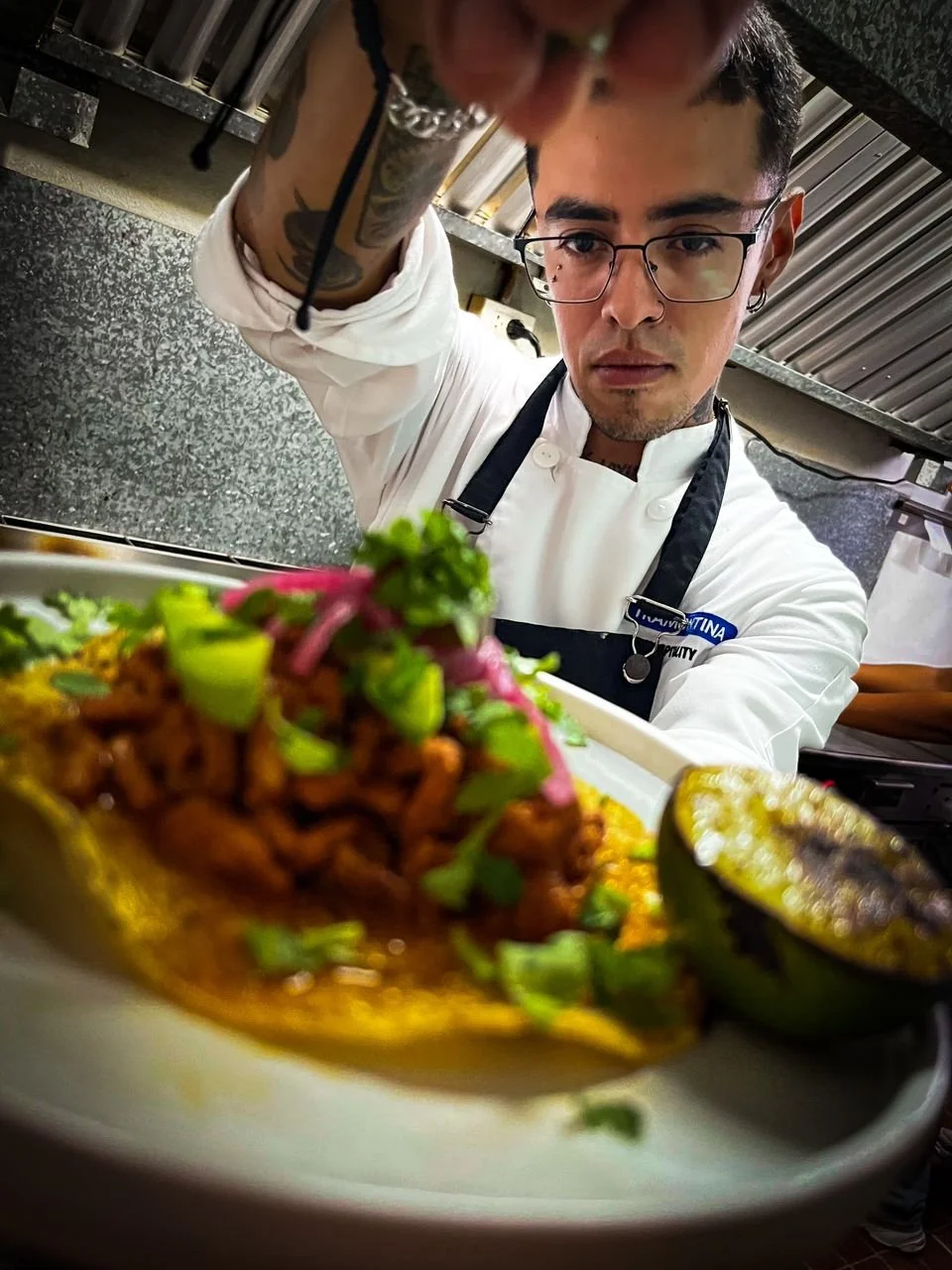 A chef garnishing a plated dish with cilantro and pink onions, with grilled lime and avocado halves nearby.