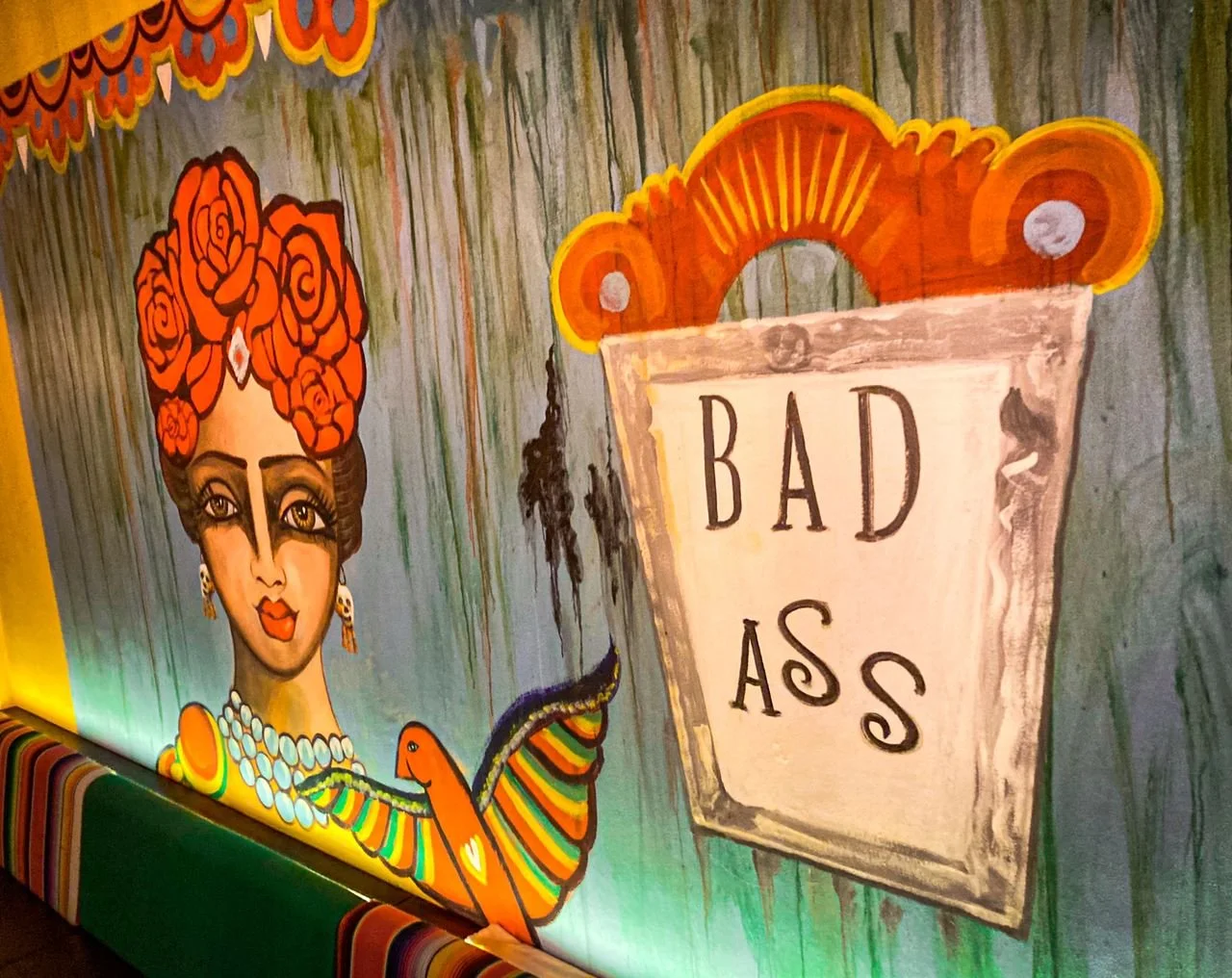 Colorful mural featuring a woman with large red roses in her hair, wearing earrings and a layered necklace, holding a snake, with a sign that reads 'BAD AS'.