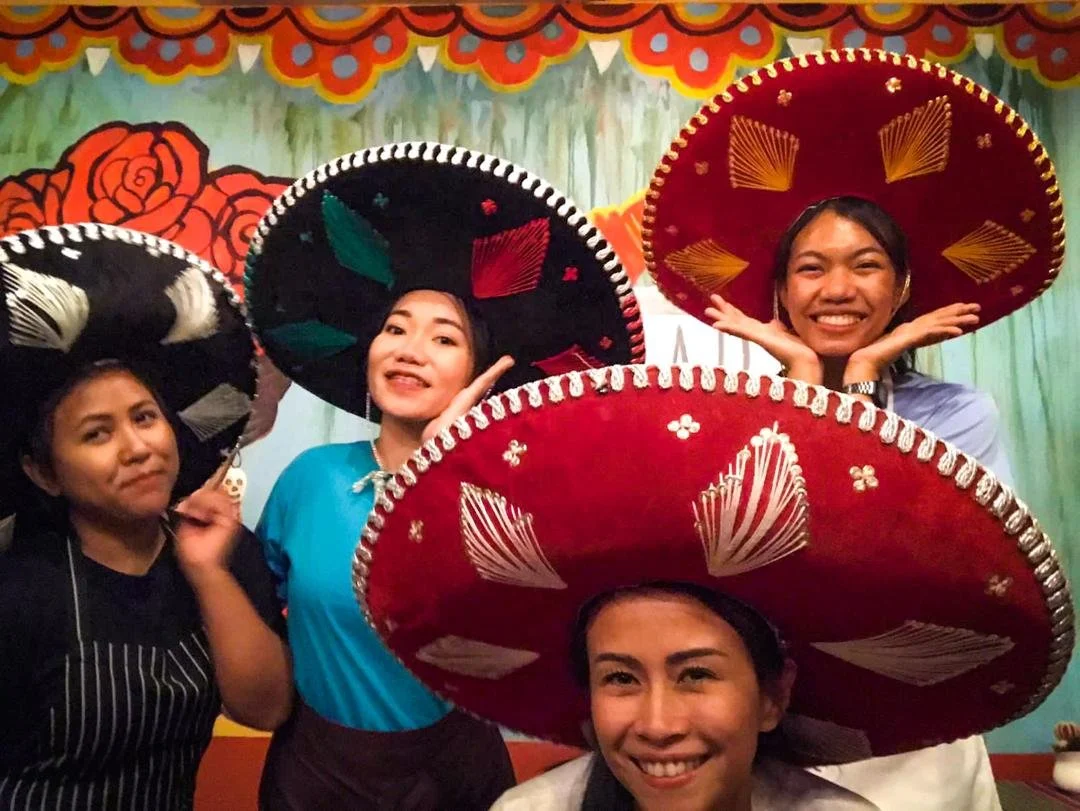 Group of four women wearing colorful sombreros and smiling, in front of a decorated backdrop with flowers.