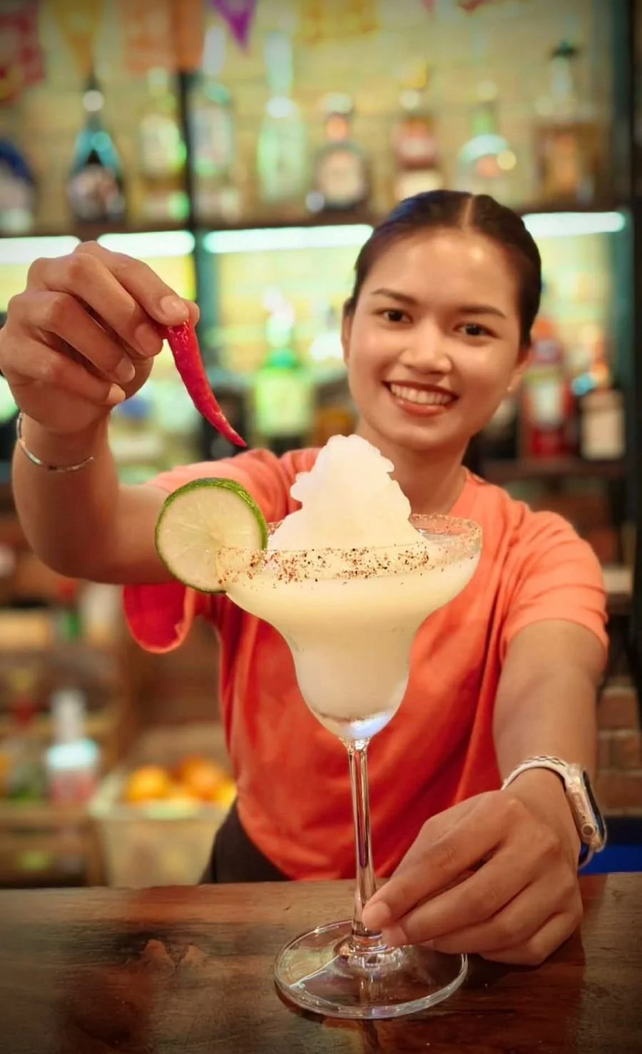A woman smiling and holding a red chili pepper over a margarita glass with lime garnish, whipped ice, and a salted rim, in a bar or restaurant setting.