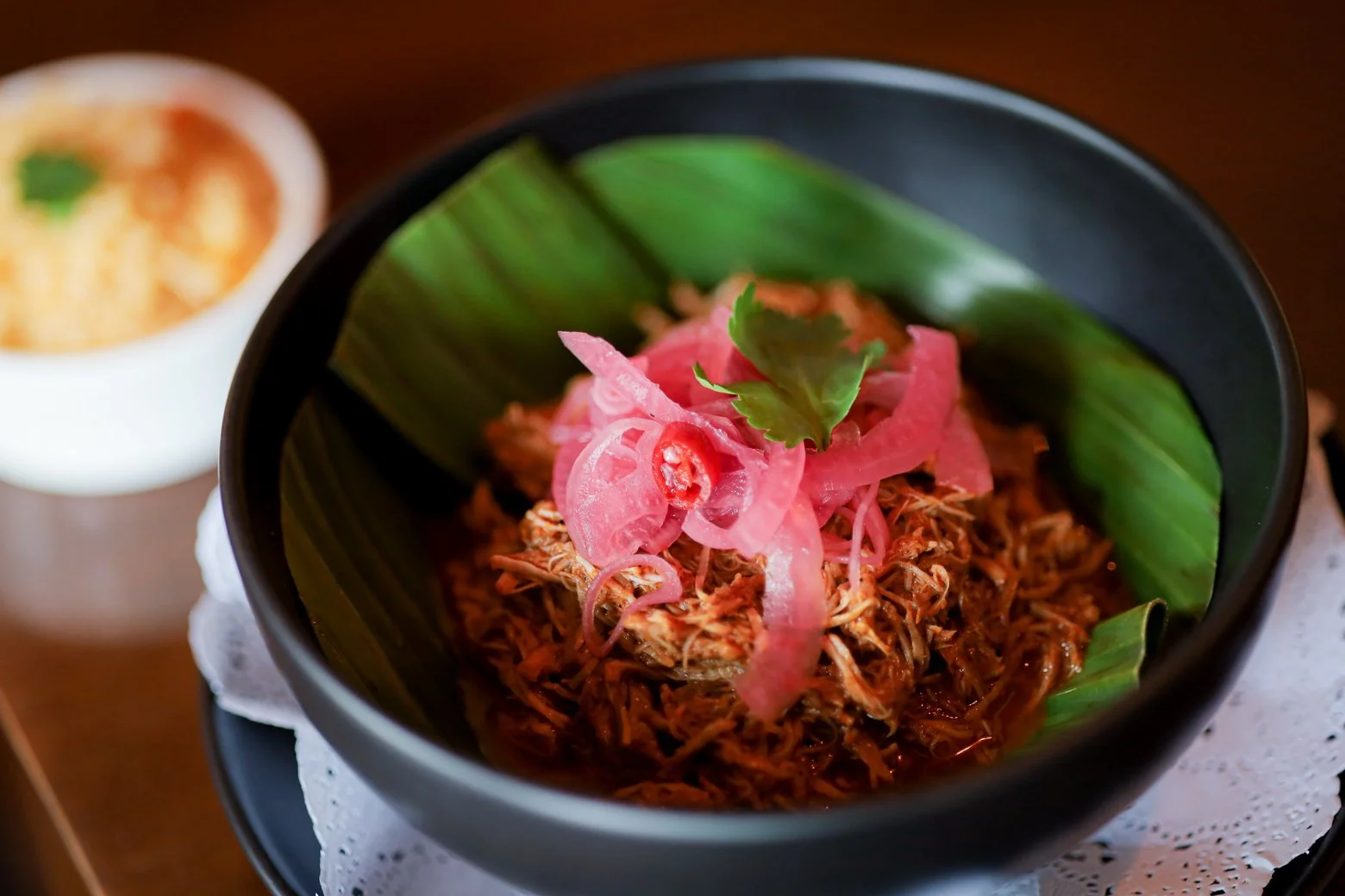Bowl of shredded meat topped with pickled pink onions and cilantro, garnished with large green leaf on the side.