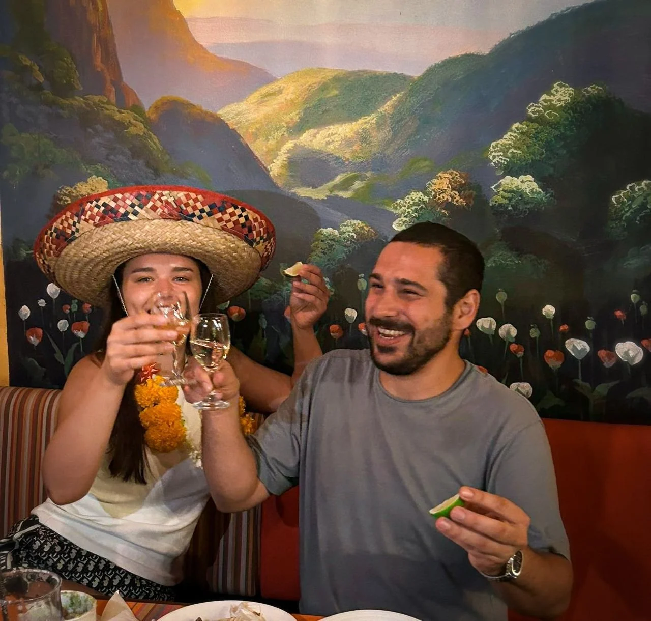 Celebrating friends in a Mexican restaurant, a woman wearing a large sombrero and flower necklace holds a shot glass and a man holds a lime slice, both smiling and toasting, with festive decoration and a mural of mountains and flowers in the backgrou