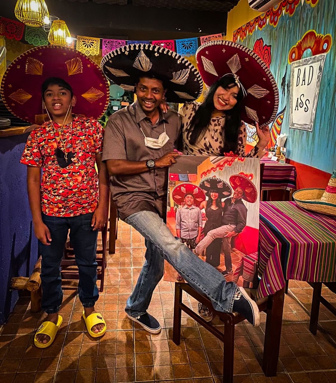 A group of three people in a festive restaurant wearing large sombreros, standing next to a colorful wall with a sign that says 'Bad Ass.' The individuals are smiling, laughing, and holding a framed photo of themselves wearing sombreros. Decor includ