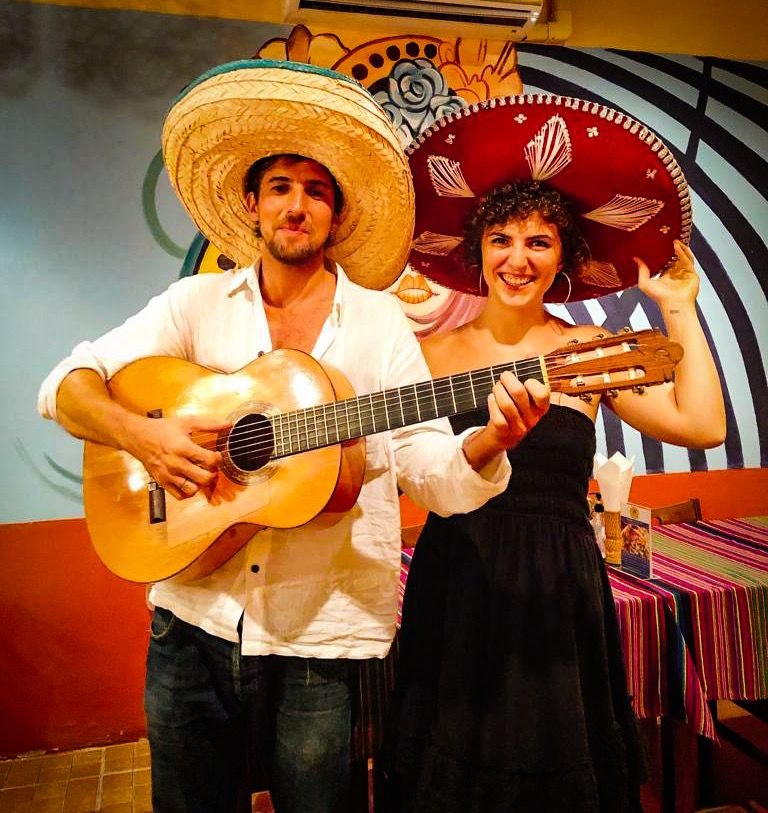 A man and woman wearing traditional Mexican sombreros, standing together. The man is holding a guitar, and the woman is smiling and touching her sombrero. They are in a colorful restaurant or party setting with vibrant wall art and striped tablecloth