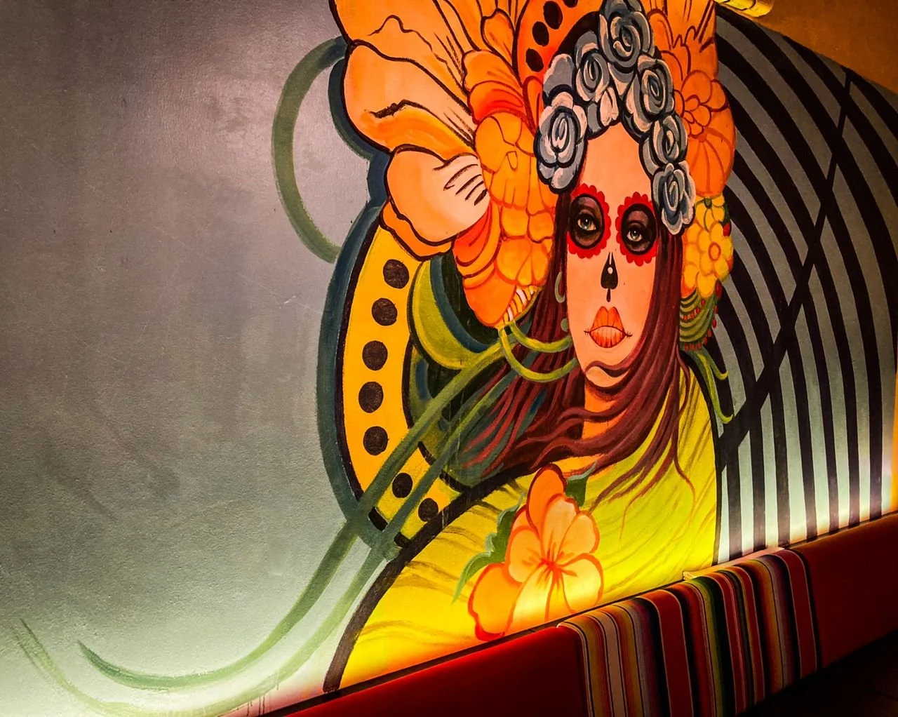 Colorful mural of a woman with sugar skull face paint, surrounded by large orange flowers, with striped patterns and a red padded bench in front.