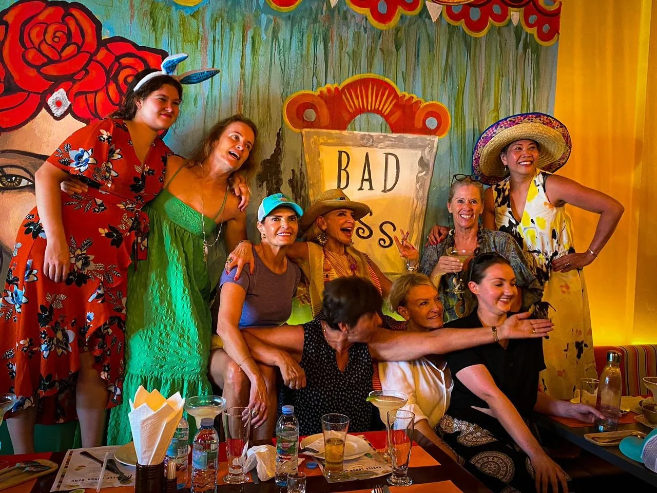 A group of women and a young girl gathered together, smiling and posing in front of a colorful painted backdrop with flowers and a sign that says 'BAD S'. Some women are wearing summer dresses and hats, and one is making a peace sign with her fingers
