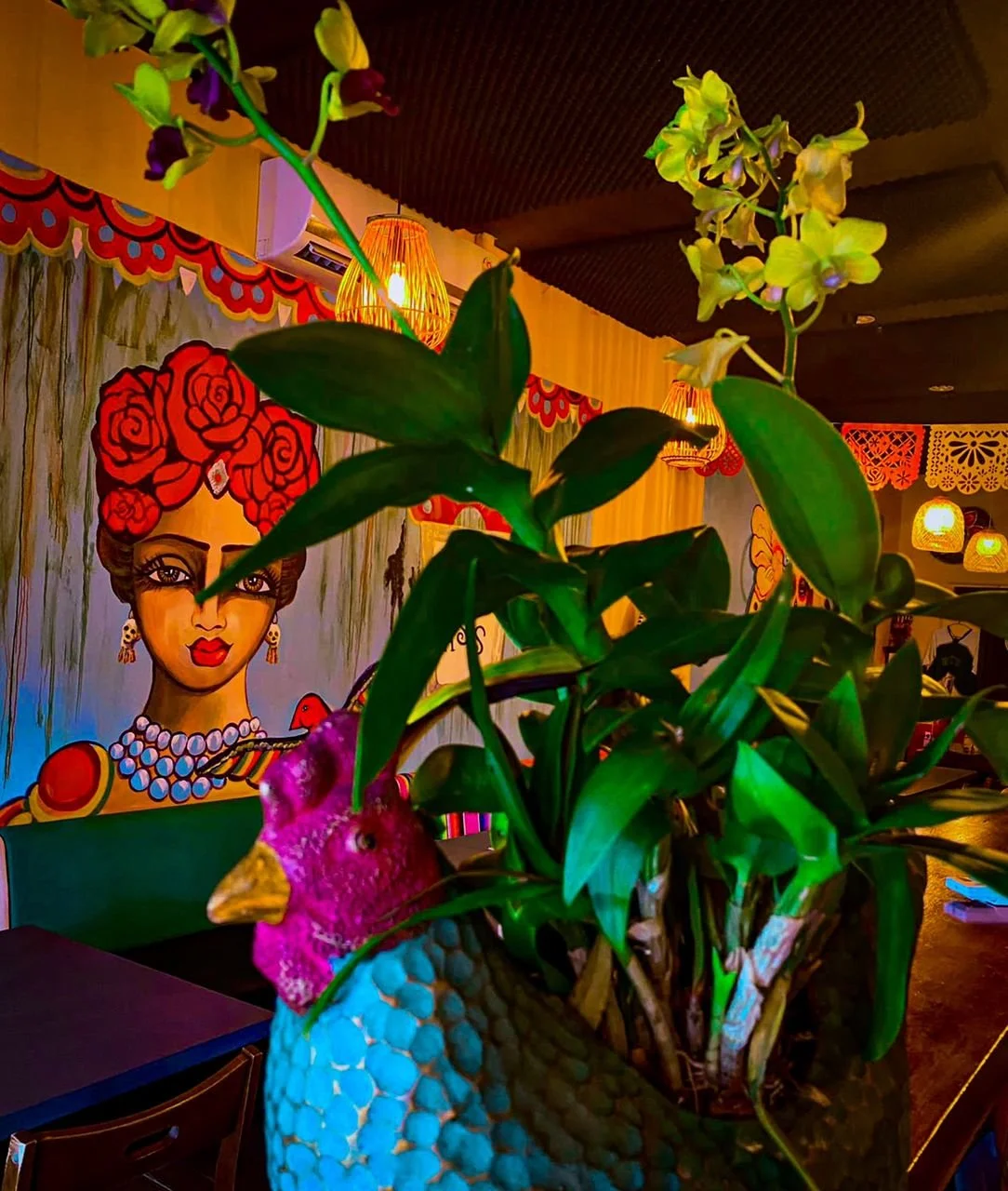 Indoor restaurant or cafe with decorative colorful mural of a woman with red flowers in her hair, wearing jewelry, and two hanging lamps. A large plant with green leaves and yellow flowers is in a decorative pot shaped like a chicken.