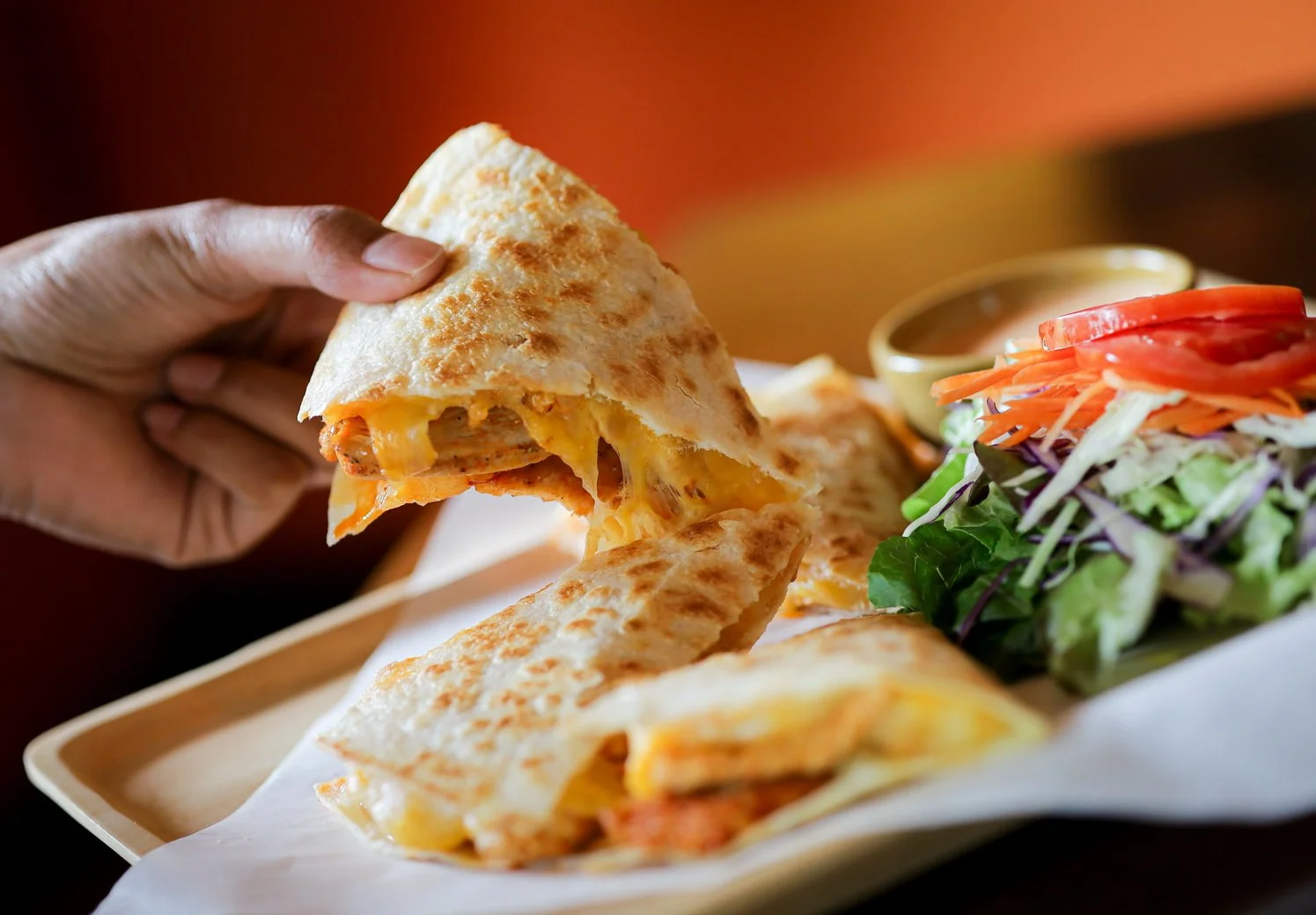 Person holding a slice of cheesy quesadilla with a side salad and dipping sauce on a tray.