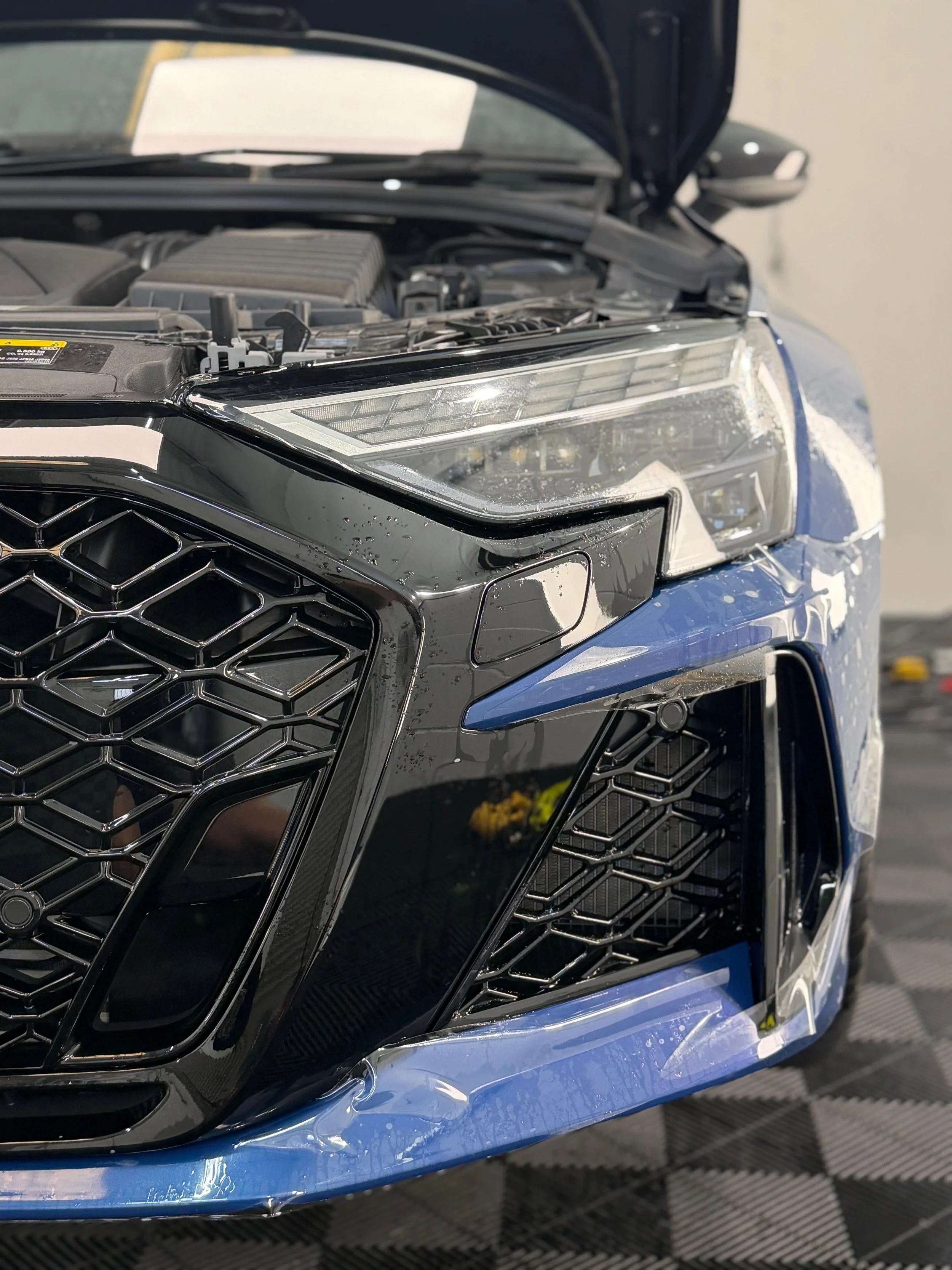 Close-up of a blue car’s front end with hood open, showcasing its grille, headlight, and XPEL paint protection film.